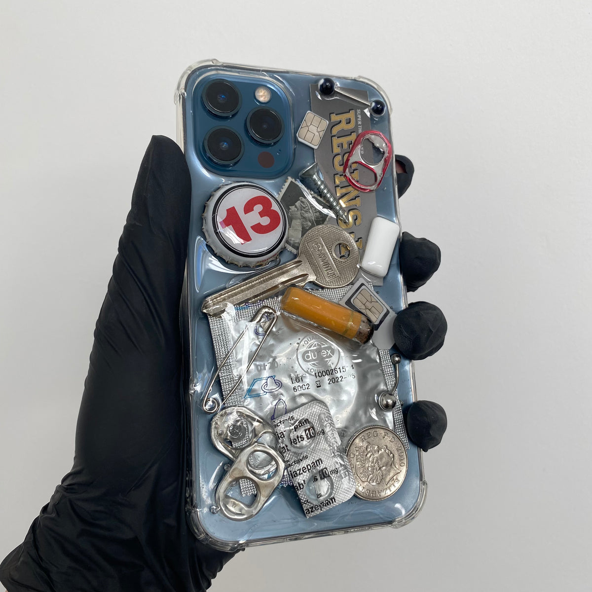 Transparent resin iPhone phone case with embedded items, including; can tabs, bottle cap, silver key, stamp, condom, safety pics, piercings, coin, gum, sim cards, screw nail, cigarette and empty pill packaging.